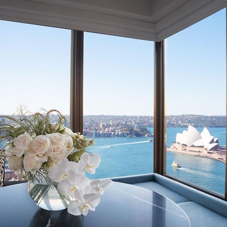 Four Seasons Hotel Sydney Esterno foto The photo shows an elegant interior space with large windows that provide a stunning view. In the foreground, there is a stylish table topped with a beautiful floral arrangement consisting of white roses and orchids in a clear vase. Through the windo