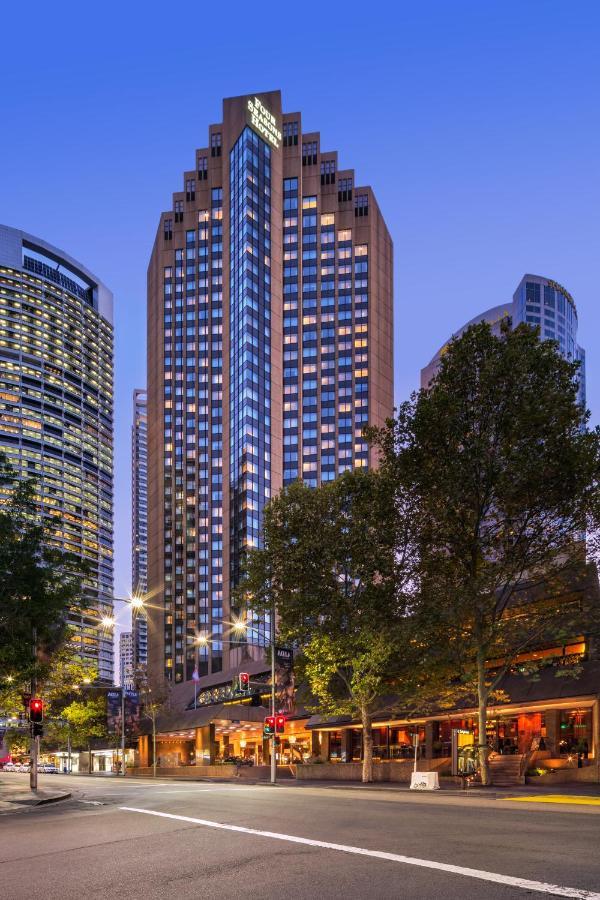 Four Seasons Hotel Sydney Esterno foto The photo shows a tall modern building with a glass façade, likely an office or residential space, illuminated in the evening. In the foreground, there are trees and streetlights, contributing to a lively urban atmosphere. Surrounding the main buildi