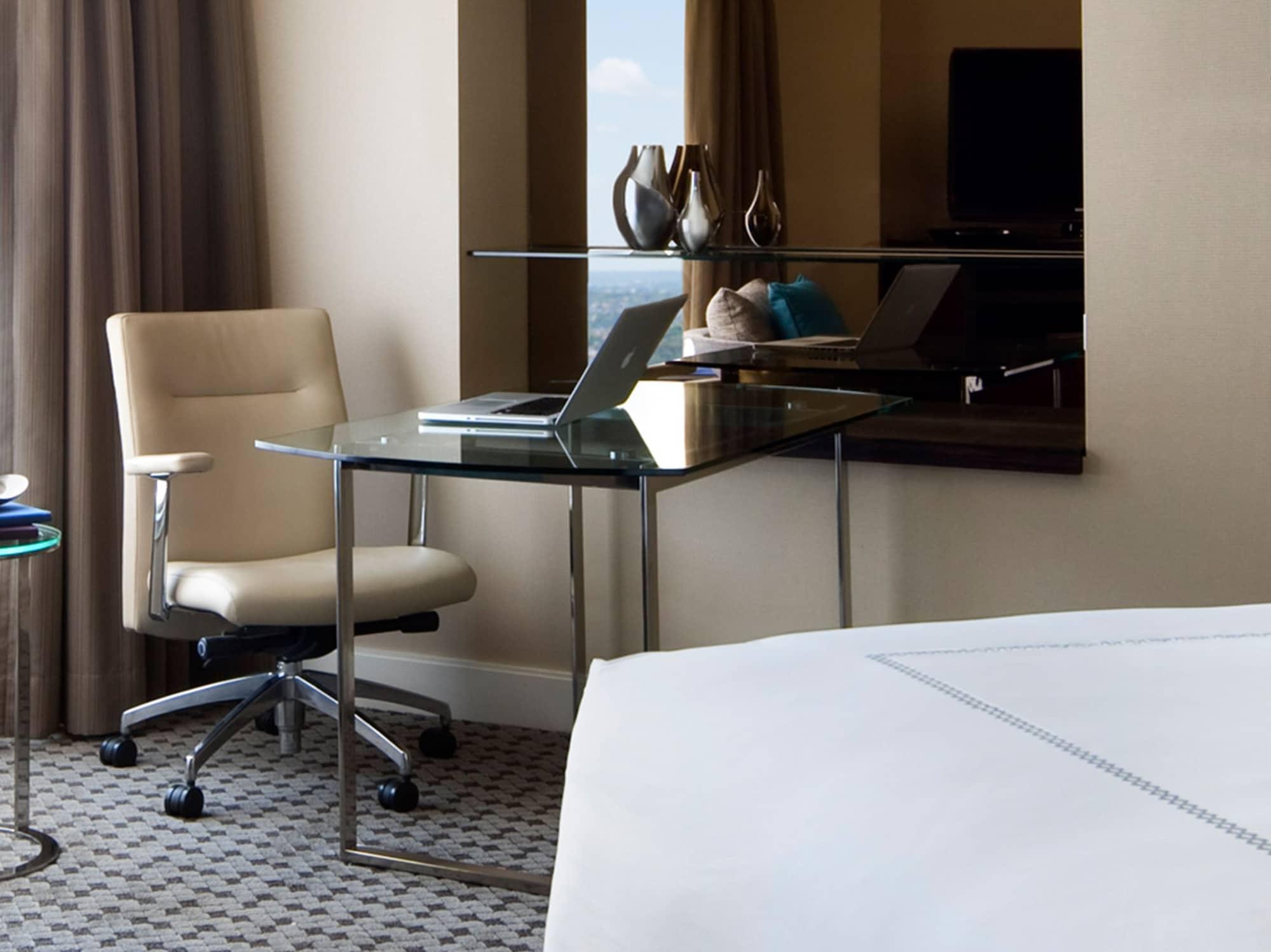 Four Seasons Hotel Sydney Esterno foto The photo depicts a modern hotel room setup. On one side, there's a sleek glass desk with a laptop set up on it, accompanied by a comfortable, ergonomic chair. The desk is positioned next to a window draped with curtains, and there are decorative ite