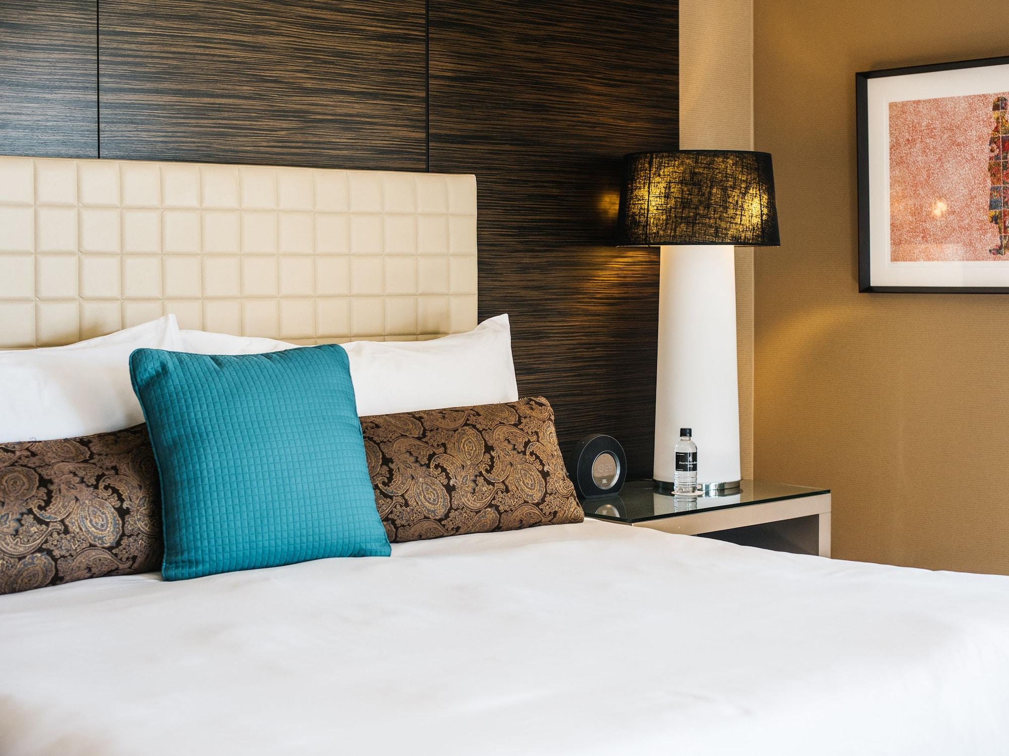 Four Seasons Hotel Sydney Esterno foto The photo shows a neatly arranged bed in a hotel room. The bed features a white duvet and a couple of decorative pillows, including a brown patterned one and a solid turquoise one. Behind the bed, there is a modern headboard with a grid pattern. Next