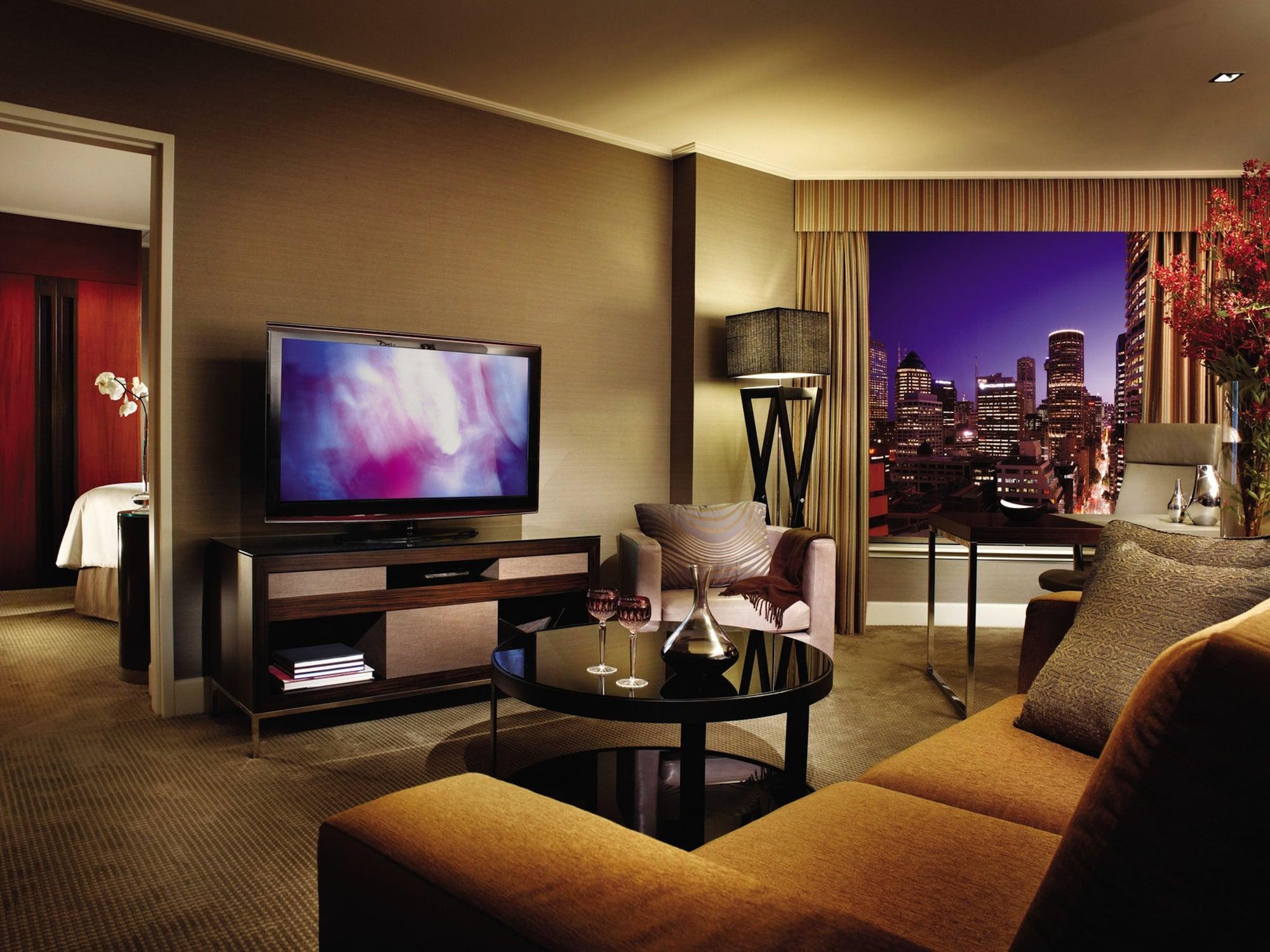 Four Seasons Hotel Sydney Camera foto The photo showcases a stylish hotel room or a modern living area. It features a comfortable brown sofa with orange cushions, a glass coffee table, and a sleek television mounted on a media console. The room is well-lit, and large windows provide a vi