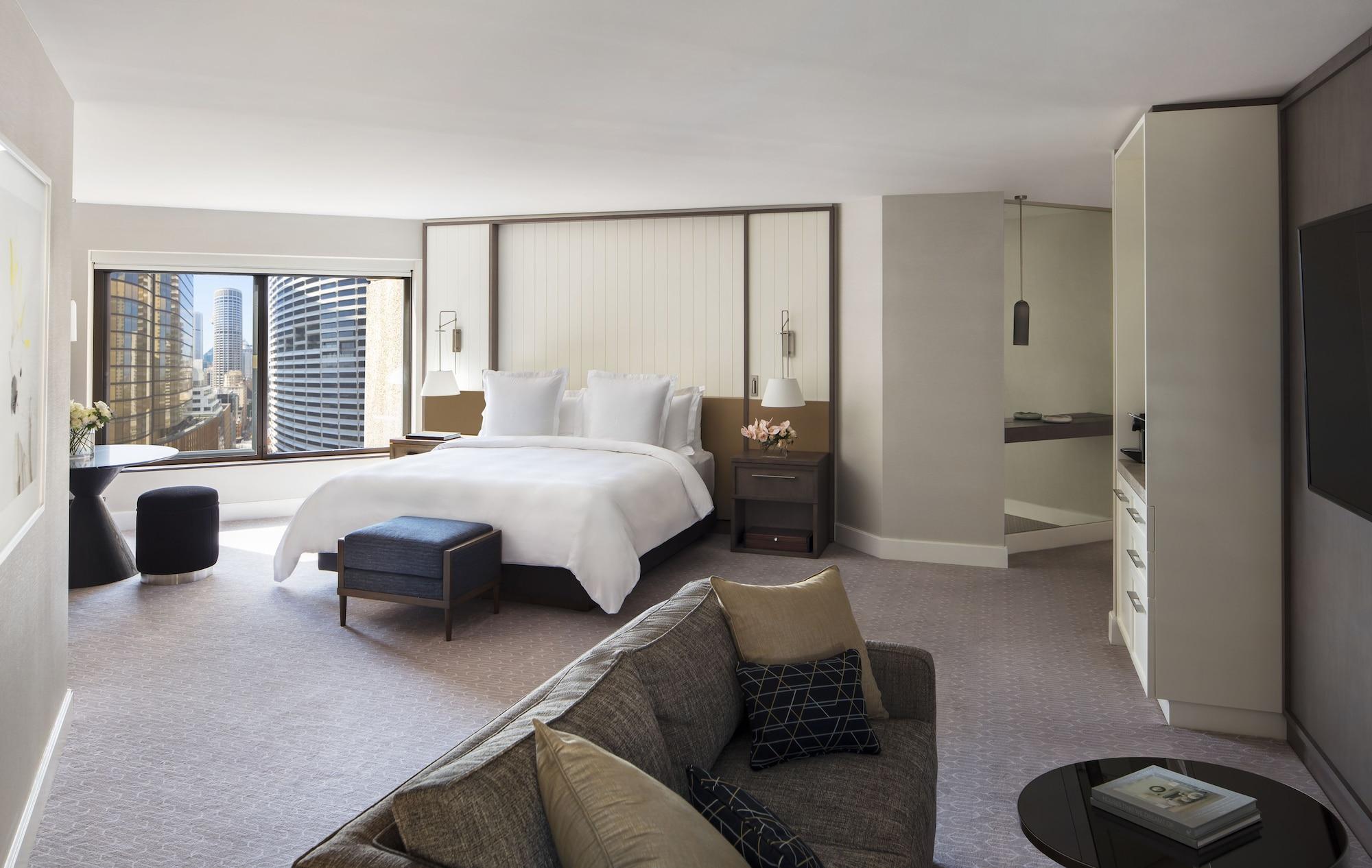 Four Seasons Hotel Sydney Esterno foto The photo shows a modern hotel room featuring a spacious layout. In the center, there's a large bed with white linens, flanked by nightstands with lamps. To one side, a comfortable sofa is positioned, complemented by a small coffee table. There is a 