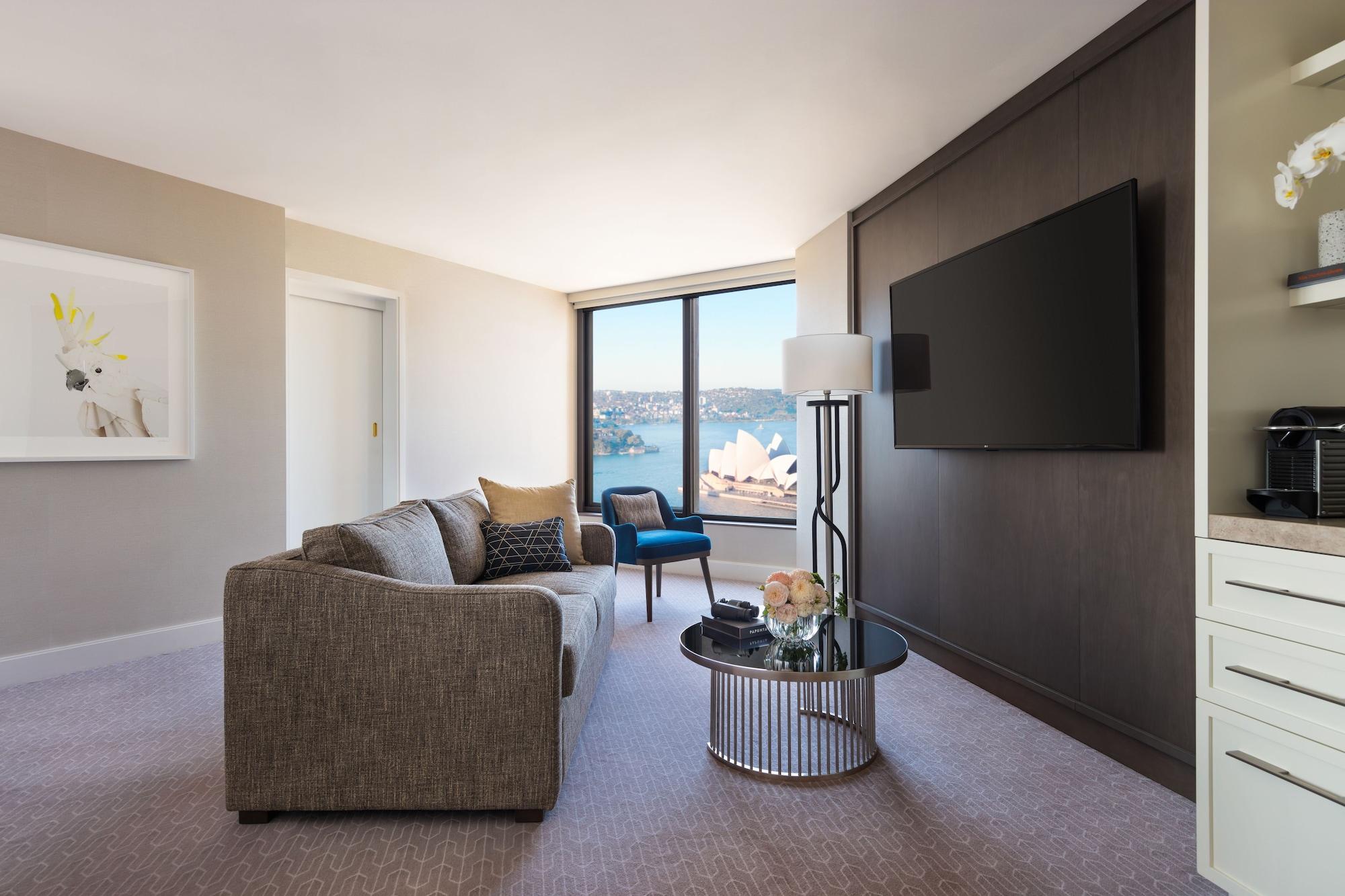 Four Seasons Hotel Sydney Esterno foto The photo shows a modern hotel room or suite. There is a comfortable gray sofa placed next to a round coffee table with a few decorative items. A tall lamp stands beside the sofa. On the opposite wall, there is a large flat-screen TV mounted, and lar