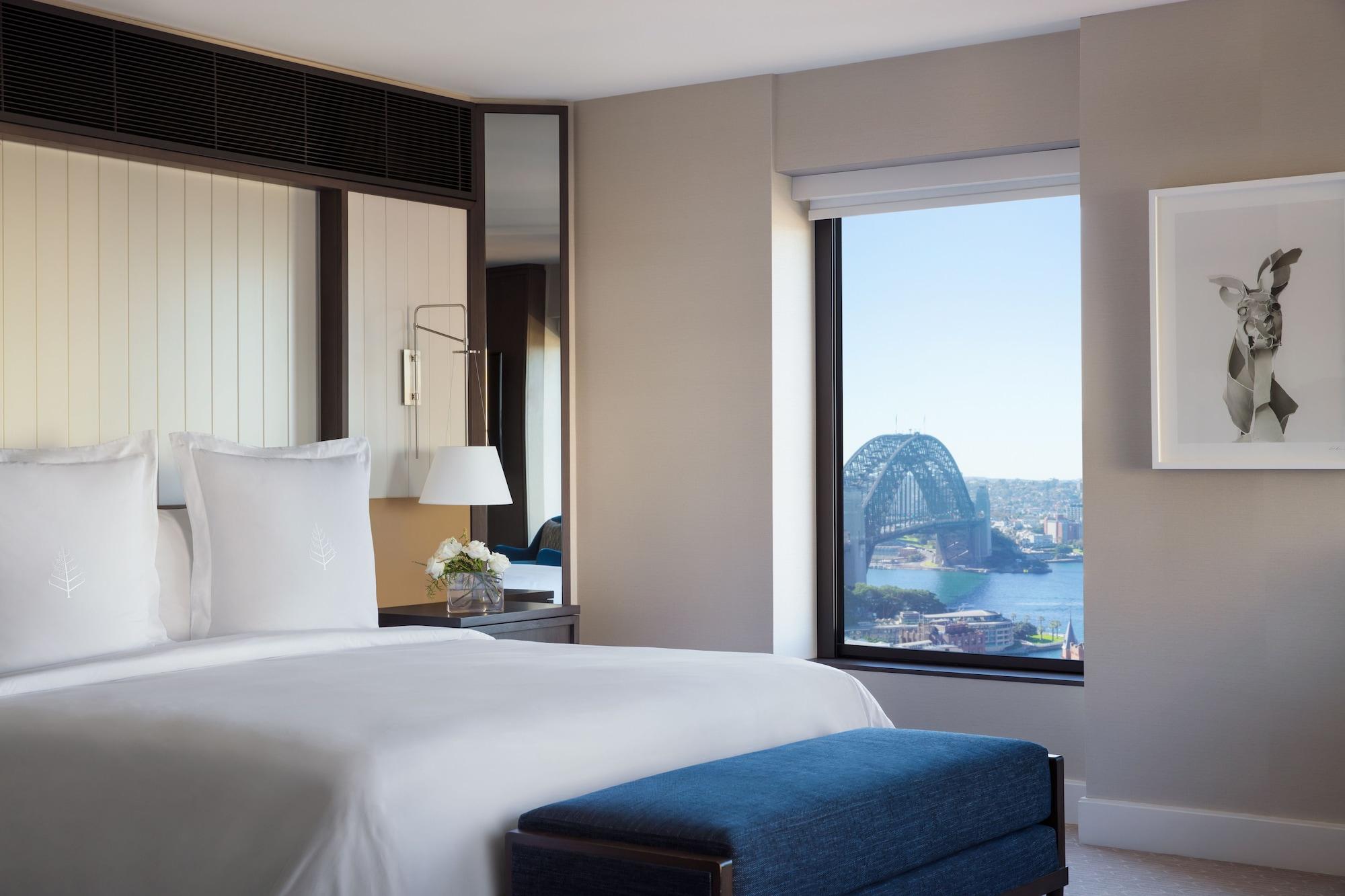 Four Seasons Hotel Sydney Esterno foto The photo shows a modern hotel room with a large bed featuring white linens and two white pillows. To the side, there is a stylish ottoman in a deep blue color. A bedside lamp and a small vase with flowers are also present. The room has a large windo