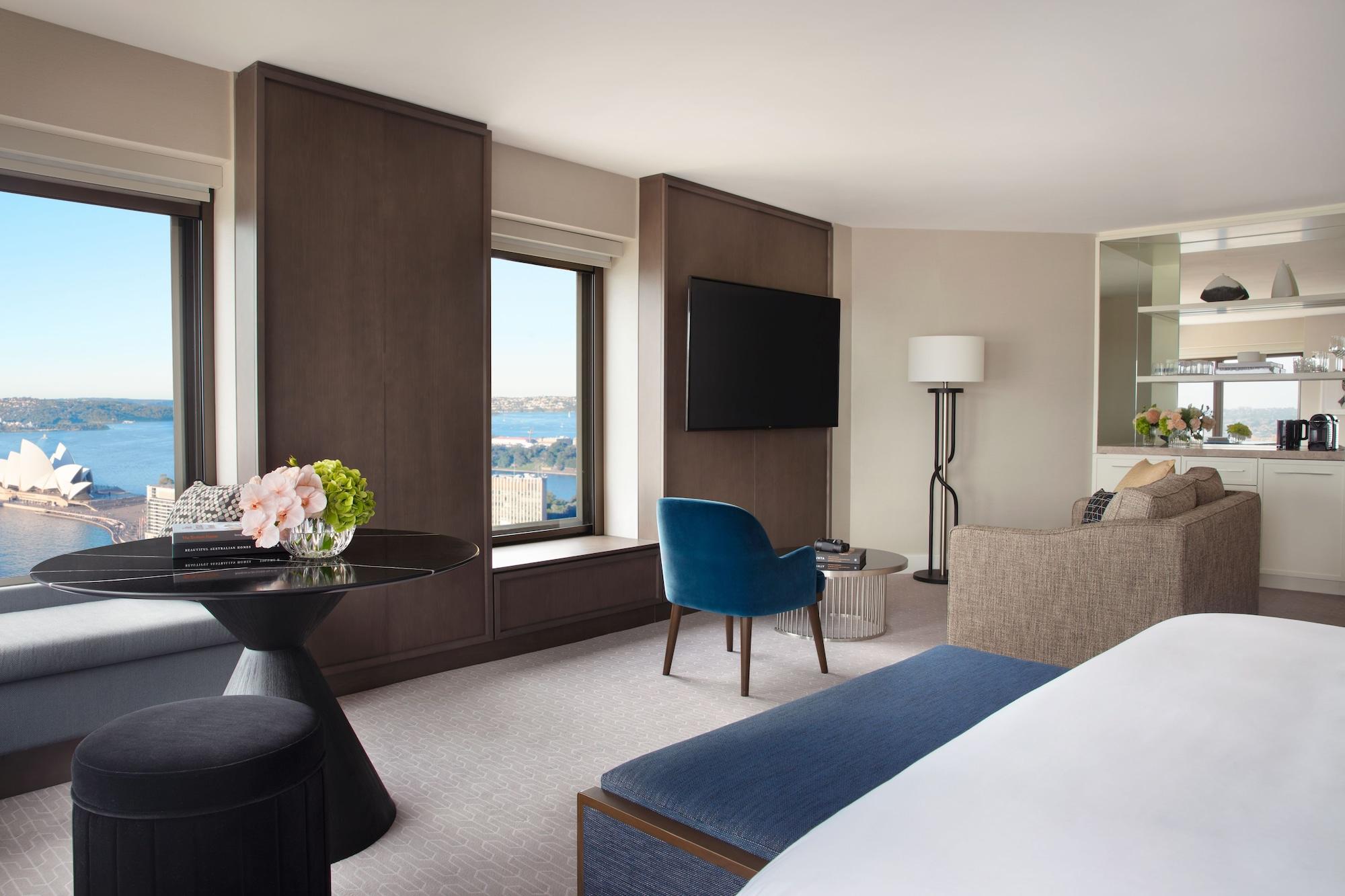 Four Seasons Hotel Sydney Esterno foto The photo shows a modern hotel room with an elegant design. It features large windows providing a view of the outside, likely overlooking a waterfront. The room includes a comfortable seating area with a beige sofa, a blue accent chair, and a small t