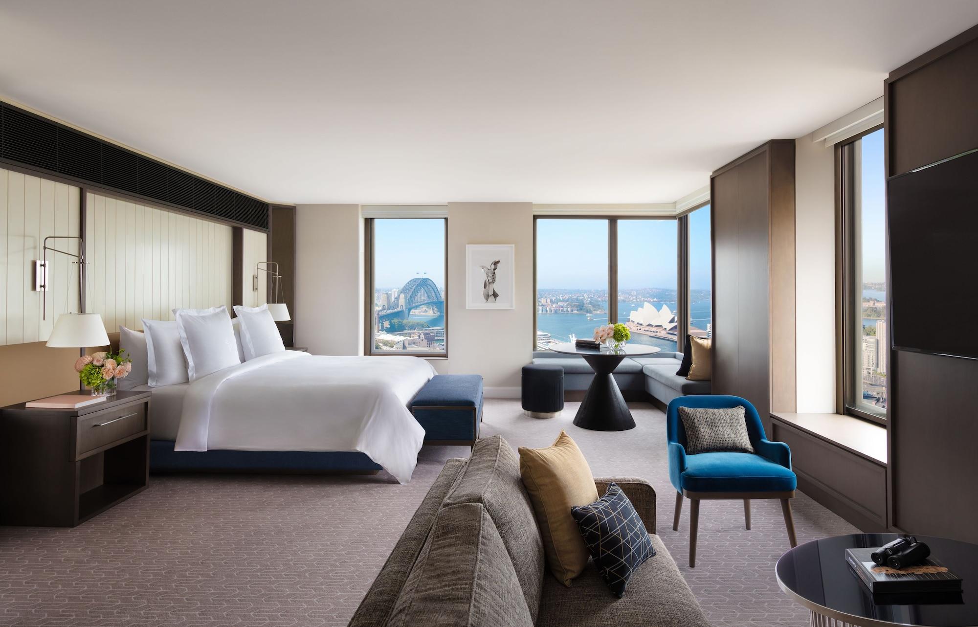 Four Seasons Hotel Sydney Esterno foto The photo shows a spacious hotel room with modern decor. It features a large bed with white linens, positioned against a wall with a contemporary headboard. There are large windows offering a view of an outdoor scene, possibly featuring a landmark li
