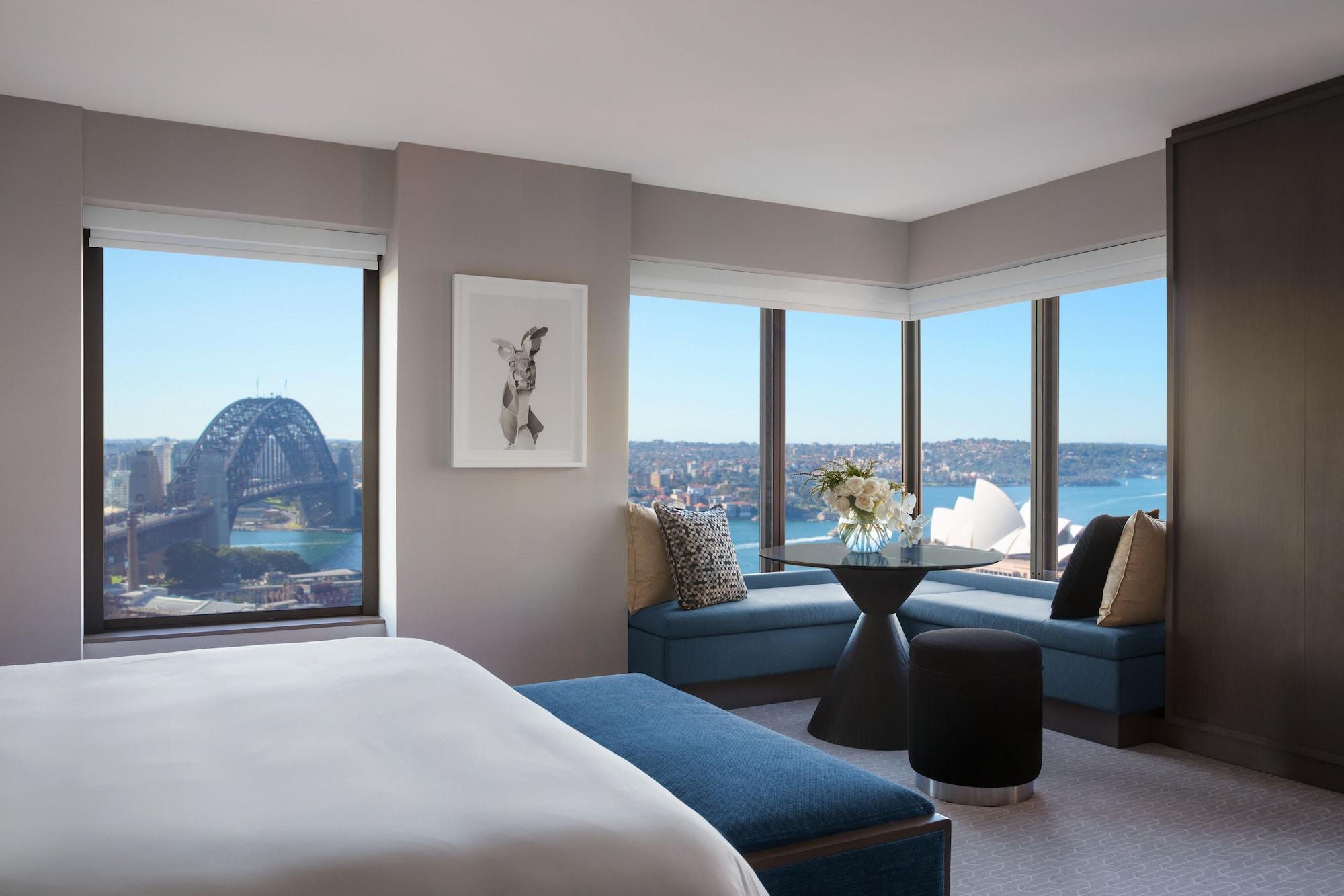 Four Seasons Hotel Sydney Esterno foto The photo shows a modern hotel room with large windows offering a panoramic view of a city skyline. In the foreground, there is a bed with white linens and a blue accent bench. A piece of artwork is displayed on the wall. In the corner, there is a ci