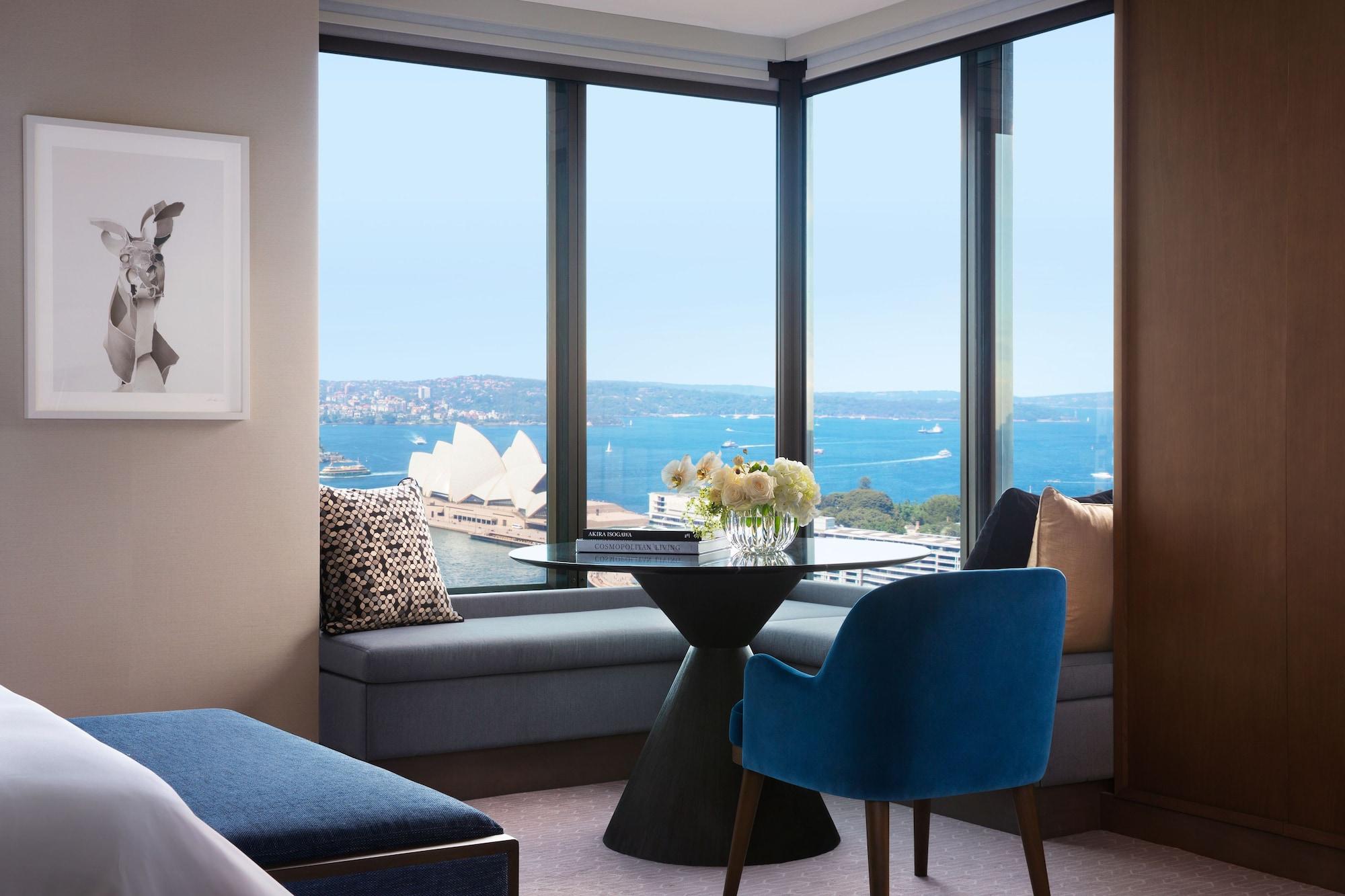 Four Seasons Hotel Sydney Esterno foto The photo shows a stylish hotel room with a large window offering a view of the water and the Sydney Opera House in the distance. There is a small, modern dining table with a vase of flowers on it, situated near the window. A comfortable blue chair i
