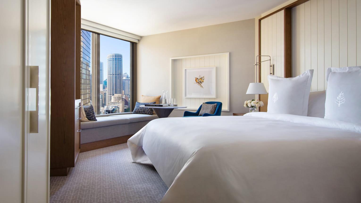 Four Seasons Hotel Sydney Esterno foto The photo shows a modern hotel room featuring a comfortable bed with crisp white linens. There is a well-lit area with a large window that offers a view of the city skyline. A cozy seating nook can be seen near the window, with a small table and chai