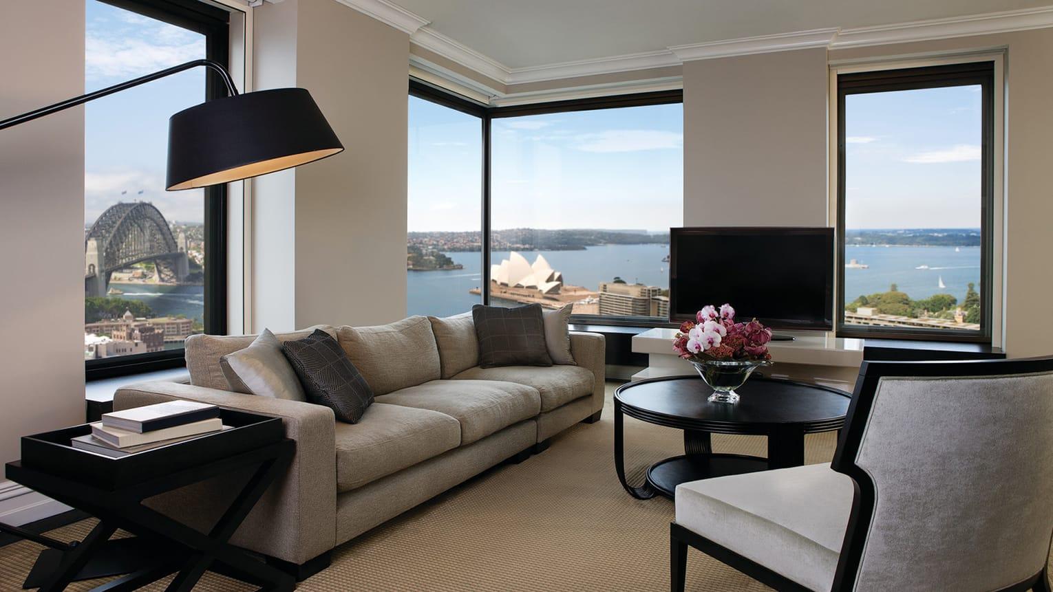 Four Seasons Hotel Sydney Esterno foto The photo shows a stylish and modern living room with large windows offering a view of a body of water and nearby buildings. The room features a comfortable, light-colored sofa and a round coffee table with a floral centerpiece. A flat-screen televis