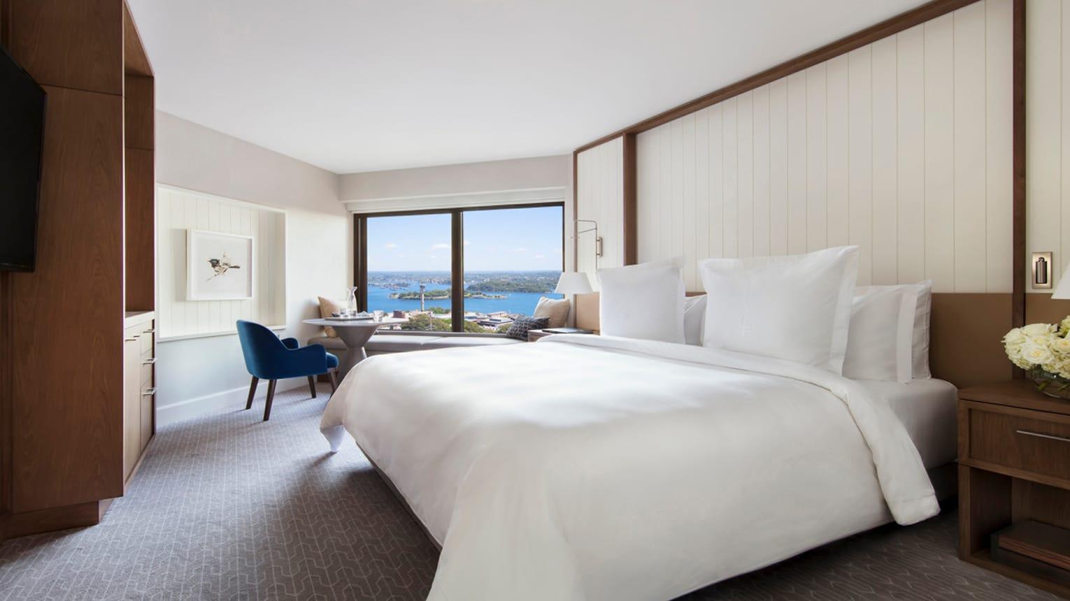 Four Seasons Hotel Sydney Esterno foto The photo shows a modern hotel room featuring a large bed with white bedding and multiple fluffy pillows. There is a window providing a view of a body of water and possibly a cityscape in the distance. Next to the bed, there's a stylish blue chair an