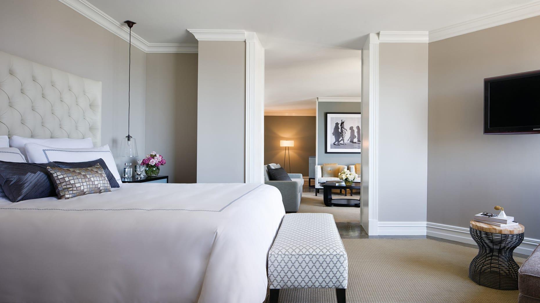 Four Seasons Hotel Sydney Esterno foto The photo depicts a stylish hotel room or suite. In the foreground, there is a neatly made bed with a light-colored comforter and decorative pillows. At the foot of the bed, there's a small bench with a patterned design. The walls are painted in neut