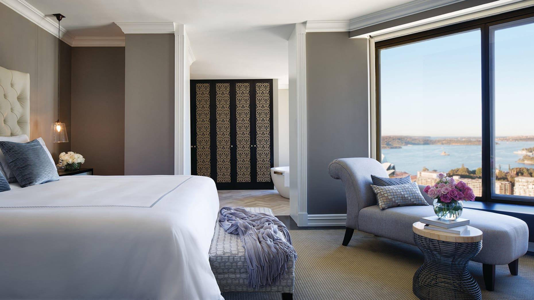 Four Seasons Hotel Sydney Esterno foto The photo shows a stylish hotel room featuring a large bed with crisp white linens. To the left, there is a comfortable seating area with a gray accent chair and a small round table topped with a vase of flowers. The room has a modern design, charact