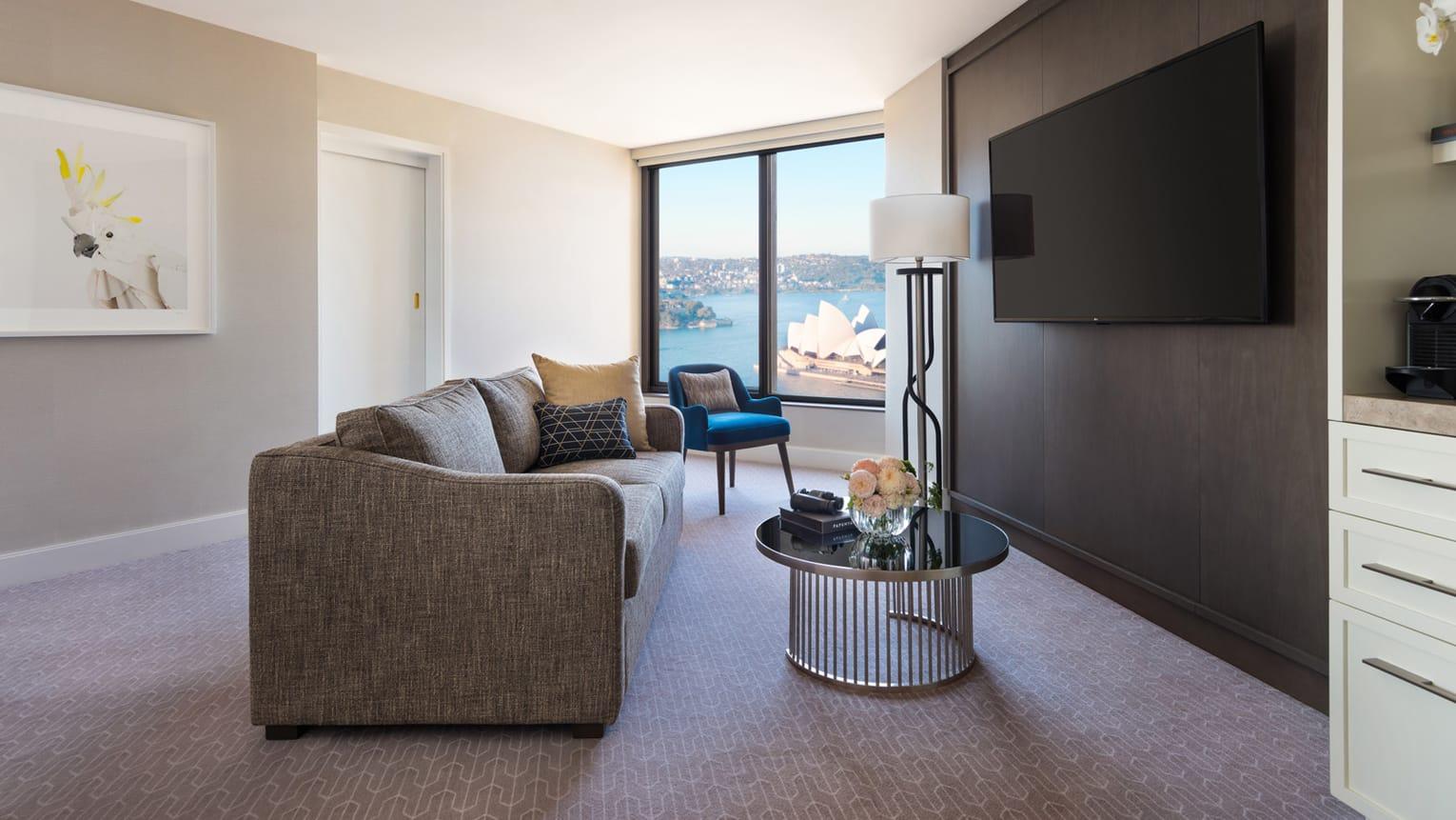 Four Seasons Hotel Sydney Esterno foto The photo shows a modern hotel room or living space. There is a comfortable gray sofa positioned in front of a large window that provides a view of the outside, likely featuring a scenic landmark. Next to the sofa, there is a small round table with a