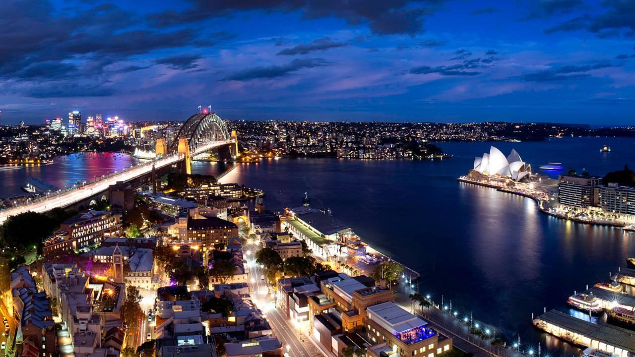 Four Seasons Hotel Sydney Esterno foto The photo depicts a stunning nighttime view of Sydney, Australia. In the foreground, you can see a bustling urban area with buildings and streets illuminated by lights. To the left, the iconic Sydney Harbour Bridge is visible, elegantly arching over 