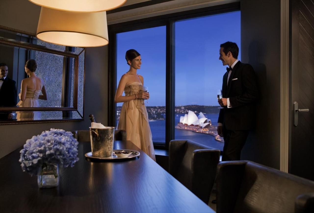 Four Seasons Hotel Sydney Esterno foto The photo depicts a stylish evening scene where a woman in an elegant dress and a man in a formal suit are engaged in conversation near a large window. They are in a modern interior space, with a table that has a bottle of champagne and a glass. The 
