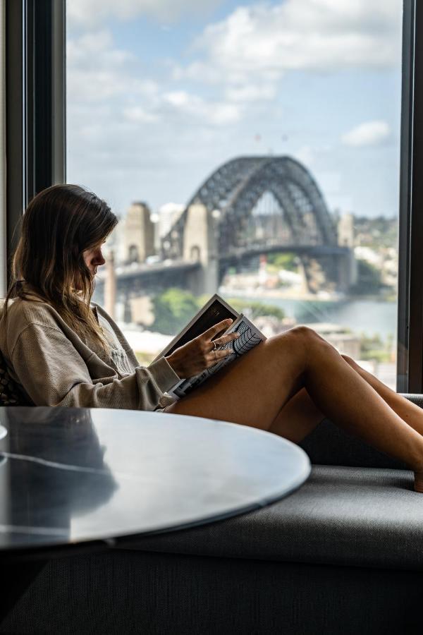 Four Seasons Hotel Sydney Esterno foto The photo depicts a woman sitting comfortably by a window, reading a book. She is wearing a casual oversized sweatshirt and is relaxed, with her legs crossed. The background features a view of a bridge, likely the Sydney Harbour Bridge, with a citysc