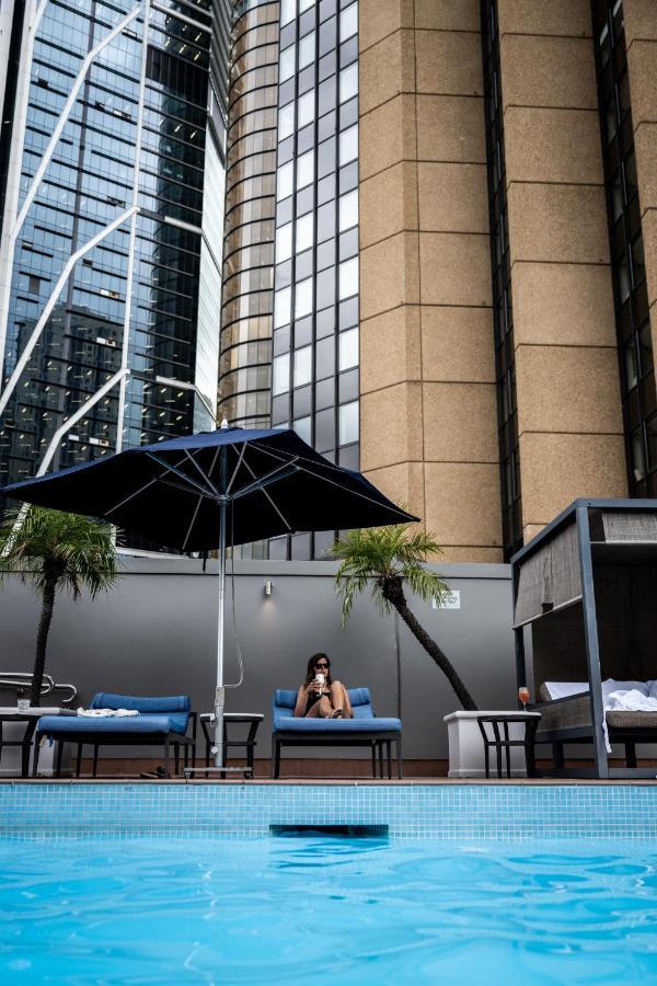Four Seasons Hotel Sydney Esterno foto The photo shows an outdoor pool area with lounge chairs under a blue umbrella. In the foreground, a person is seated on one of the lounge chairs, looking at a phone. There are palm trees nearby, and the background features tall buildings with a mix o