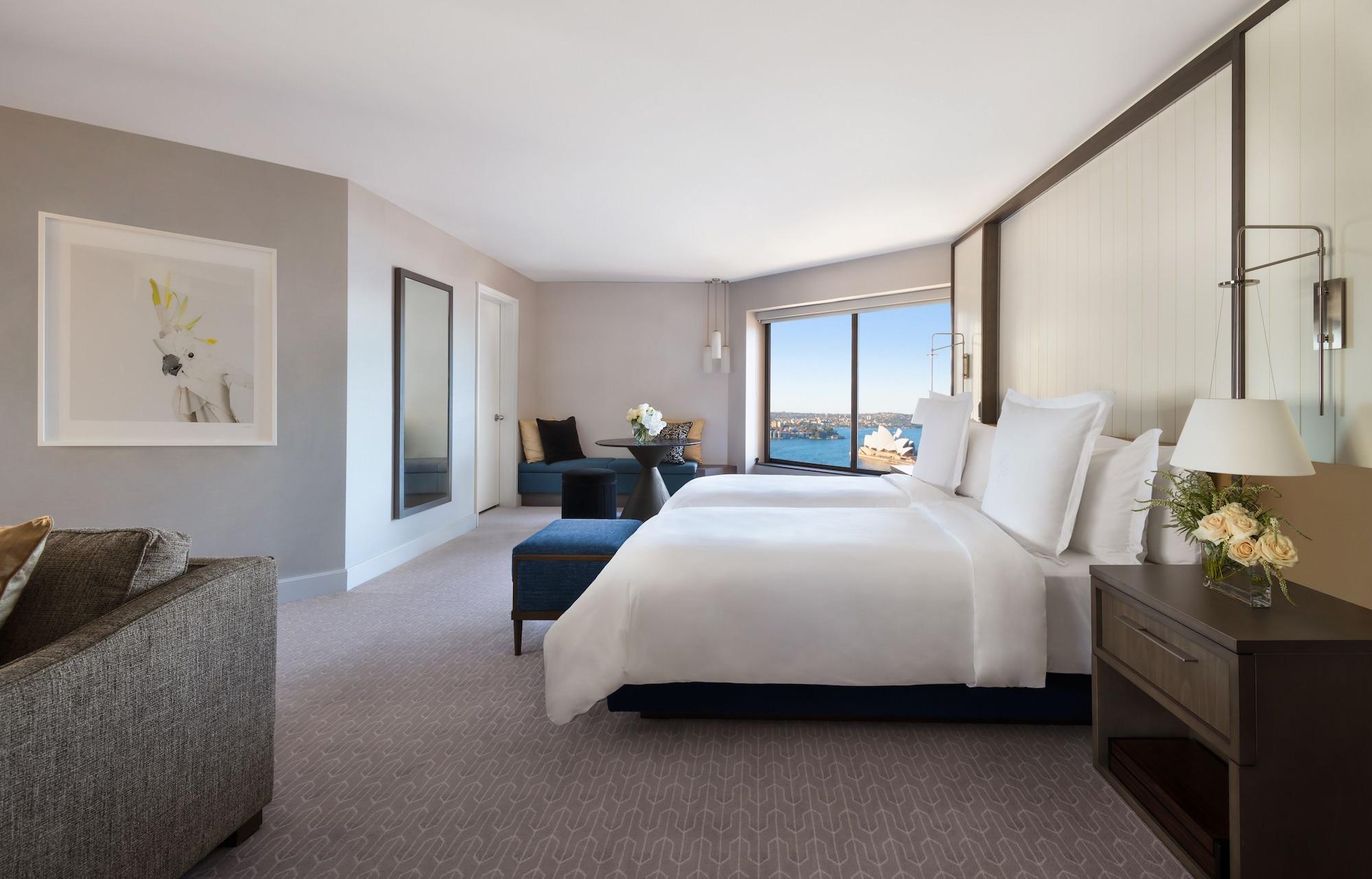 Four Seasons Hotel Sydney Esterno foto The photo shows a modern hotel room with a clean and elegant design. It features two large beds with white linens and plush pillows, positioned near a large window that offers a view of a body of water. In the room, there is also a comfortable seatin