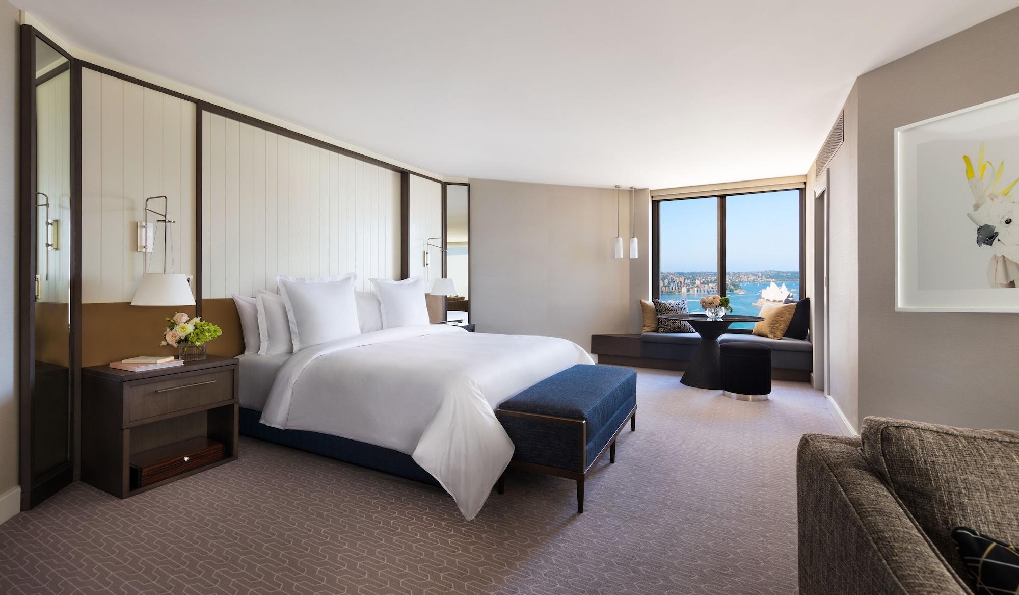 Four Seasons Hotel Sydney Esterno foto The photo shows a modern hotel room that features a spacious layout. In the center, there is a large bed with white linens, complemented by a blue upholstered bench at the foot of the bed. The walls have a light and neutral color scheme, creating a c