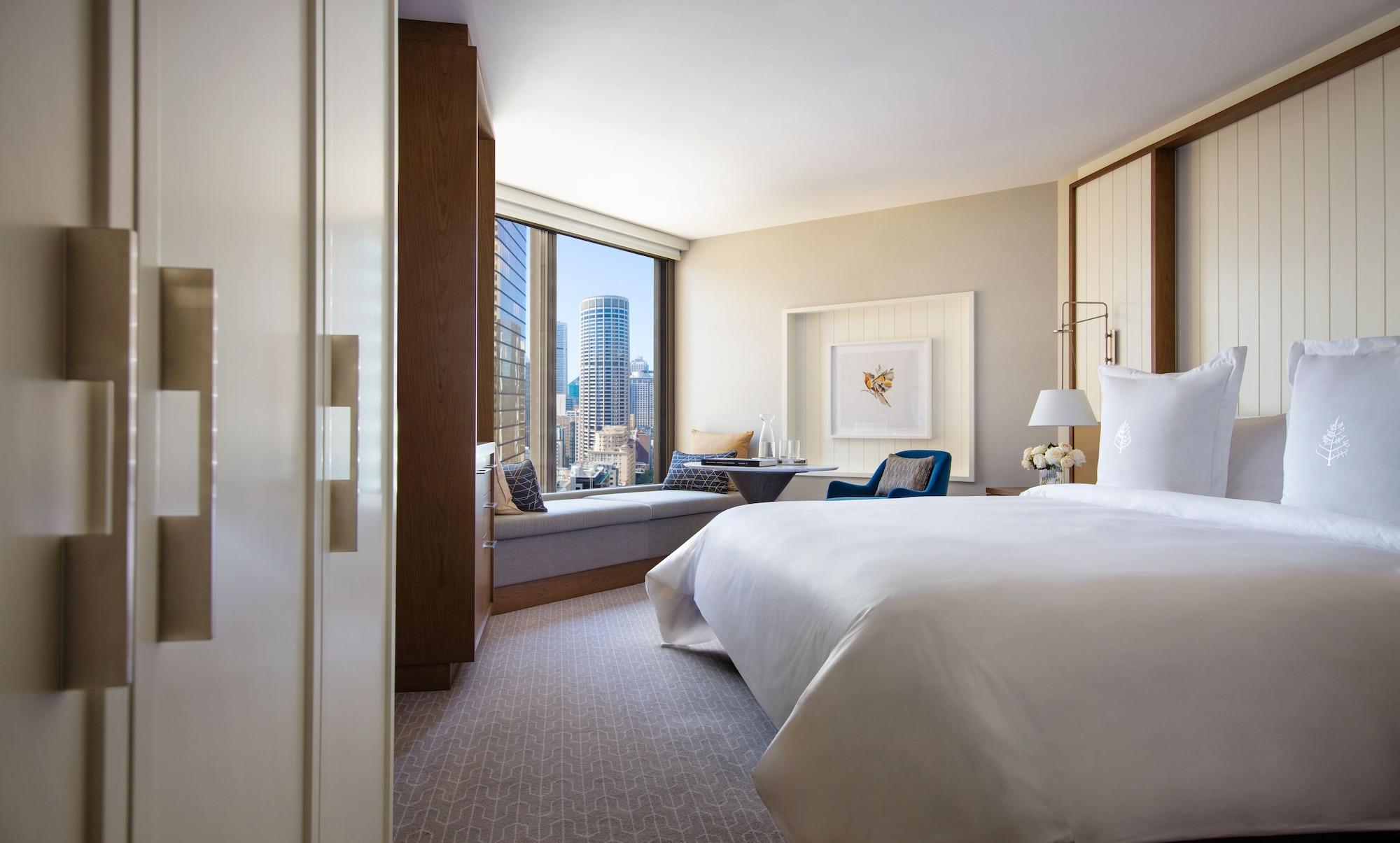 Four Seasons Hotel Sydney Esterno foto The photo shows a modern hotel room with a large bed, featuring a crisp white duvet and pillows. There is a stylish seating area with a cozy window nook and comfortable chairs, allowing for a view of the city skyline outside. The room has contemporar