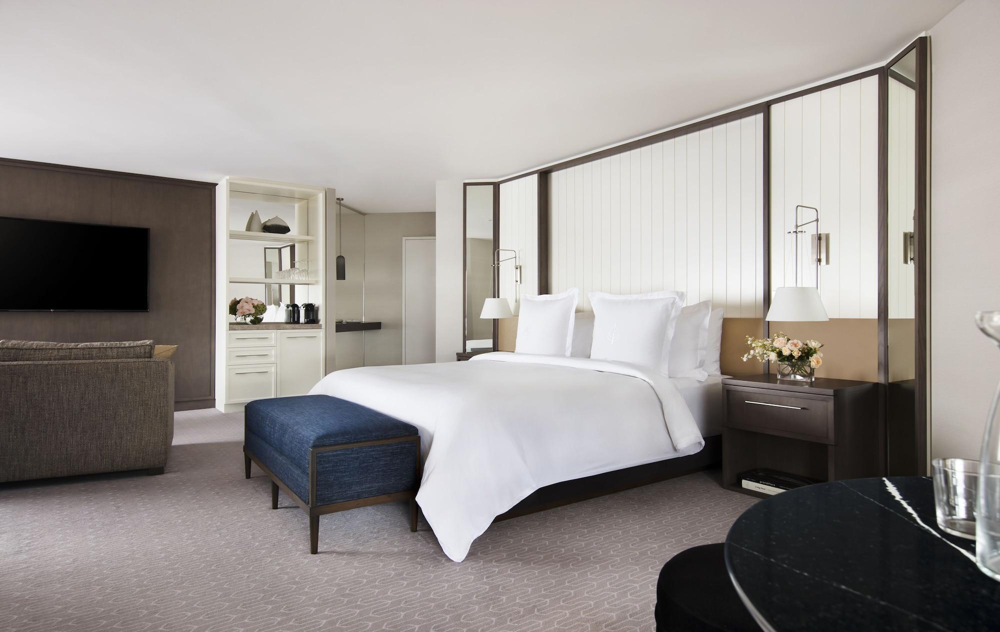 Four Seasons Hotel Sydney Esterno foto The photo depicts a modern and elegantly designed hotel room. It features a spacious layout with a large bed covered in white bedding, flanked by bedside tables and lamps. There is a stylish blue ottoman at the foot of the bed, adding a touch of colo