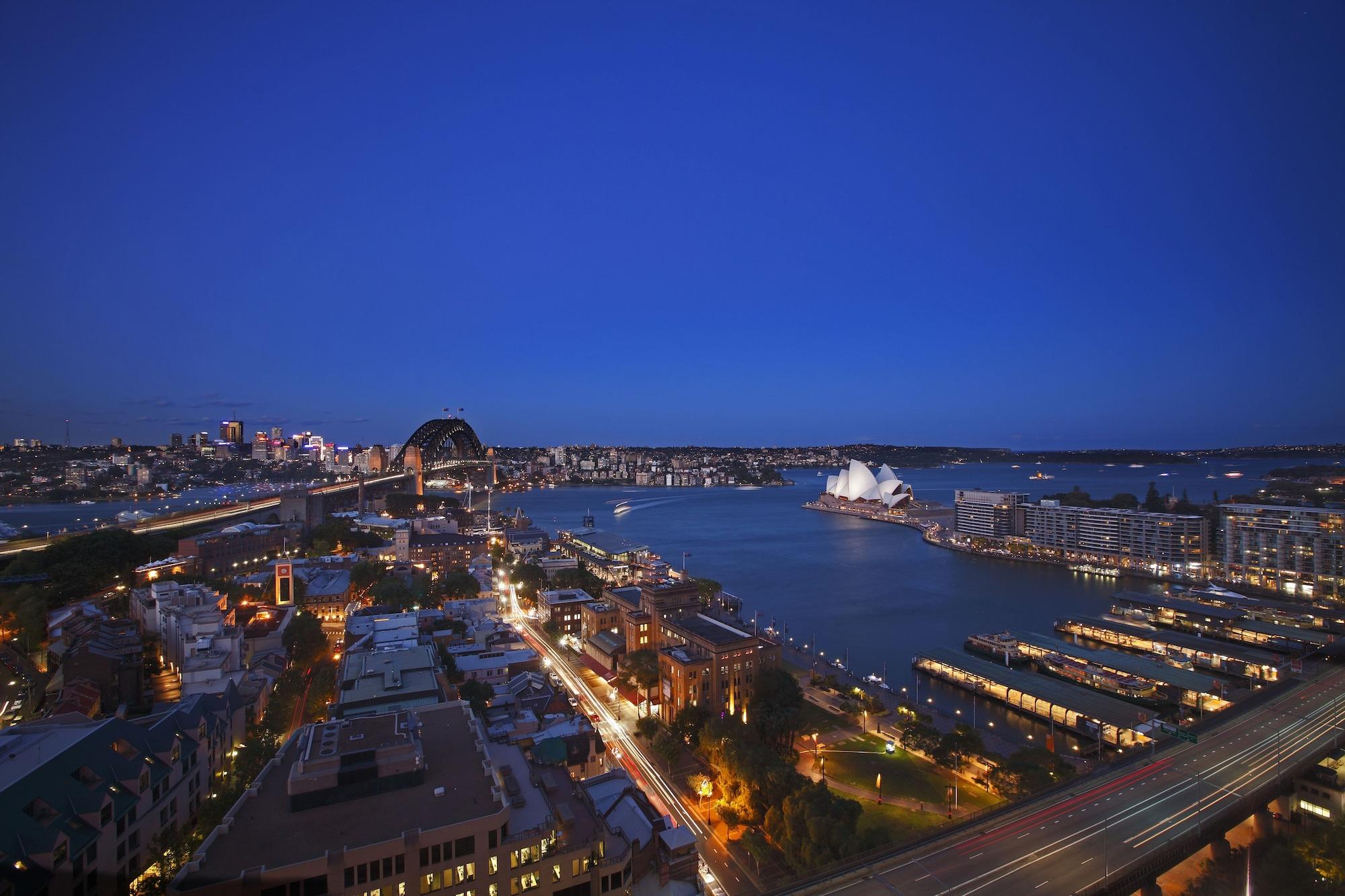 Four Seasons Hotel Sydney Esterno foto The photo captures a scenic view of Sydney, Australia, during twilight. The iconic Sydney Opera House is prominently visible along the waterfront, characterized by its distinctive white sail-like roof. In the background, the silhouette of the Sydney 