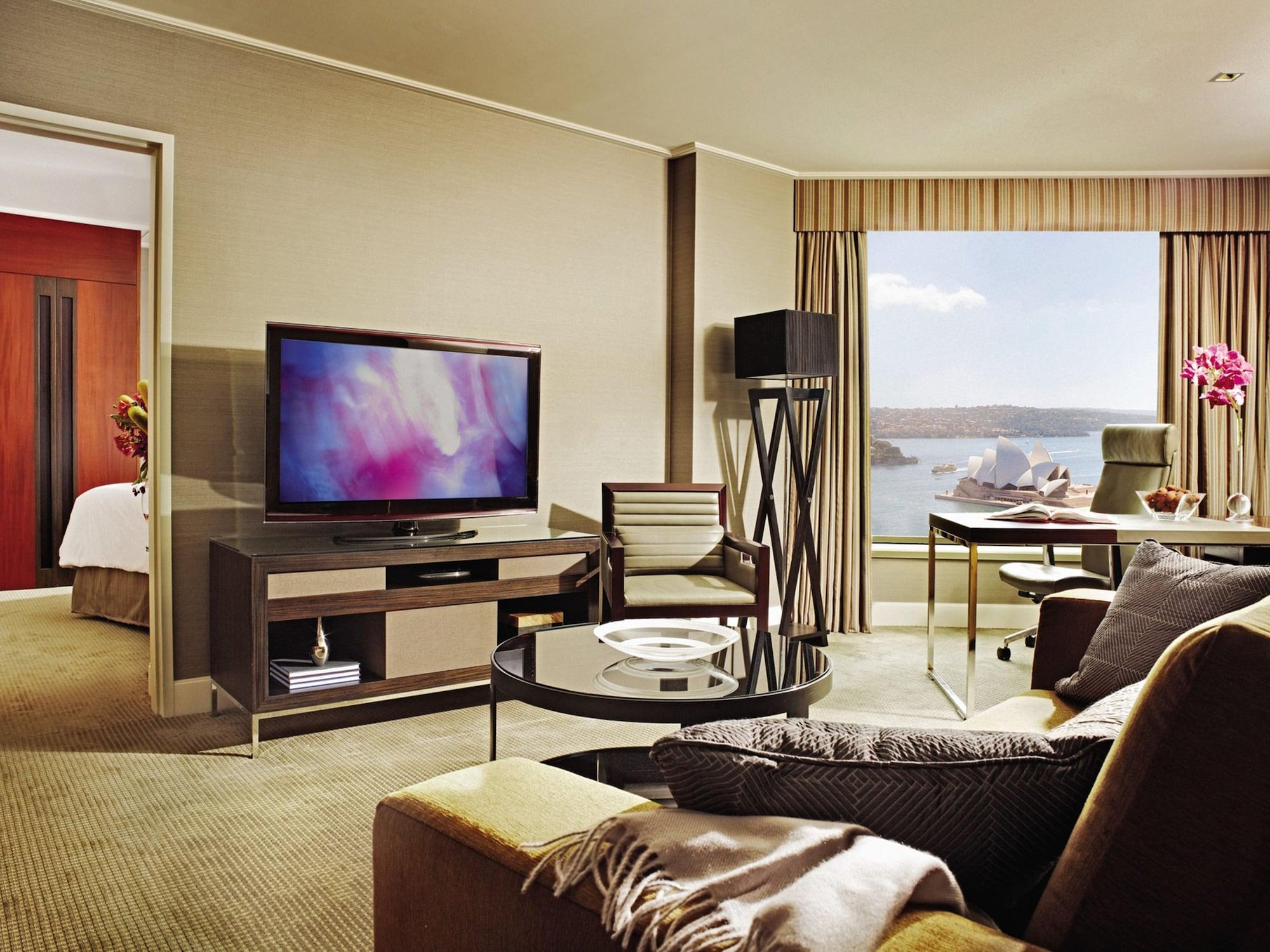 Four Seasons Hotel Sydney Camera foto The photo shows a well-furnished hotel room or living space. It features a cozy seating area with a large, comfortable couch and a coffee table. A flat-screen television is mounted on a cabinet, and there is a stylish chair nearby. A desk with a chai