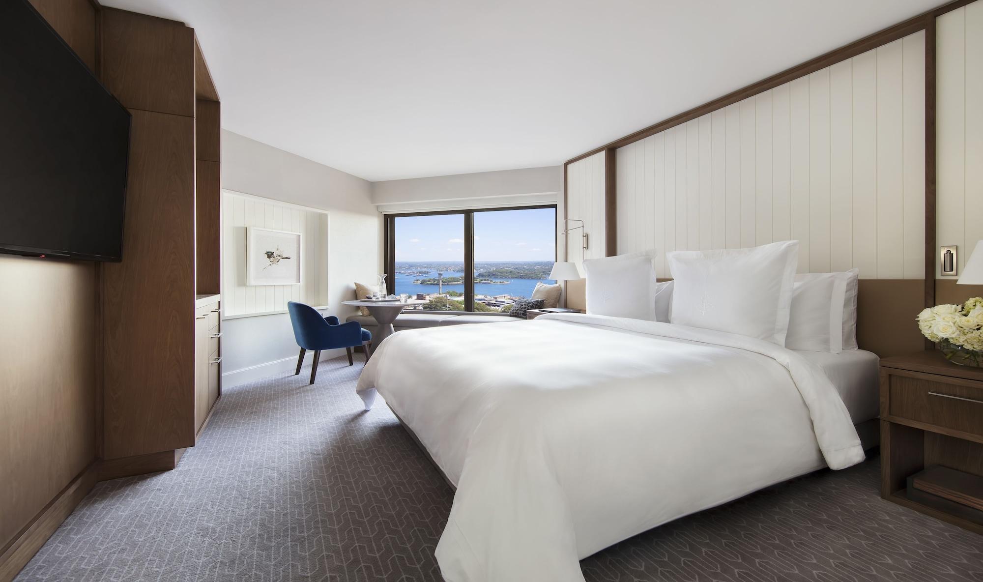 Four Seasons Hotel Sydney Esterno foto The photo shows a modern hotel room designed with a clean and elegant aesthetic. The room features a large bed with white linens and multiple pillows. There is a blue chair positioned near a window that offers a view of the outdoors, likely showcasin