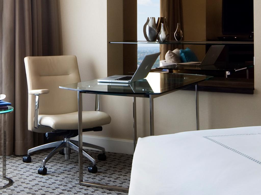 Four Seasons Hotel Sydney Esterno foto The photo shows a modern hotel room setting with a sleek, minimalist design. In the foreground, there is a glass-top desk supported by a metal frame. A comfortable beige office chair is positioned at the desk. On the desk, there is a laptop, suggesti