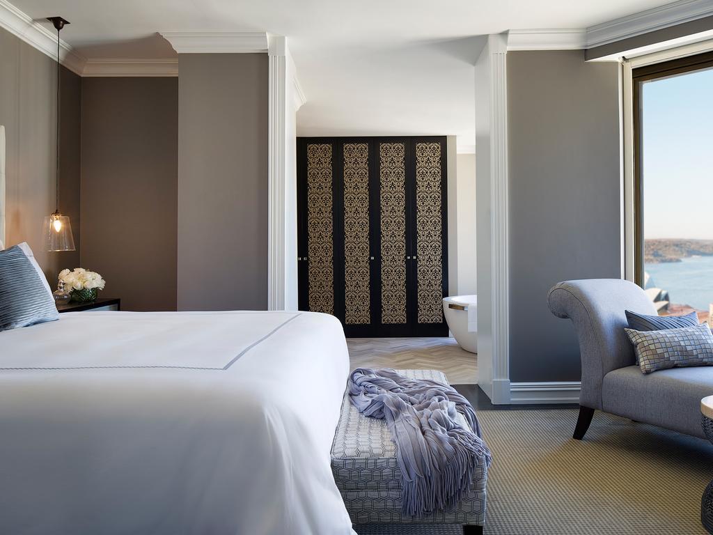 Four Seasons Hotel Sydney Esterno foto The photo shows a luxurious hotel room or suite. There is a large bed with white linens positioned towards the center. A gray armchair is nearby, accompanied by a small ottoman or stool. The walls have a soft gray tone, and there are elegant decorati