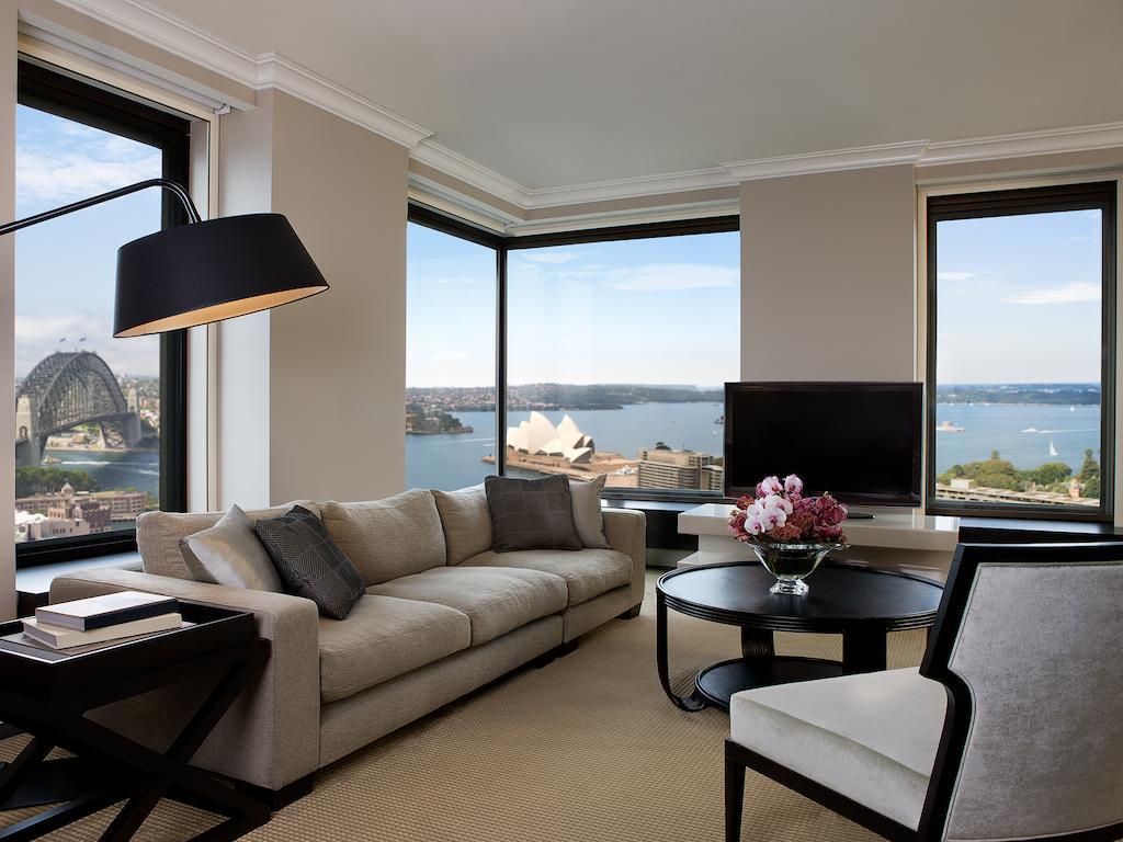 Four Seasons Hotel Sydney Esterno foto The photo depicts a modern and elegantly designed living room with large windows offering a panoramic view of a waterfront scene. The room features a light-colored sofa with decorative cushions, a round coffee table in the center, and a stylish floor