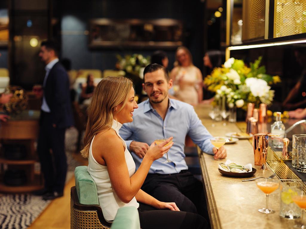 Four Seasons Hotel Sydney Esterno foto The photo depicts a stylish bar or lounge setting. In the foreground, a woman is sitting at the bar, smiling and engaging in conversation with a man who is seated beside her. She is holding a drink and wearing a sleeveless top, while the man is dress