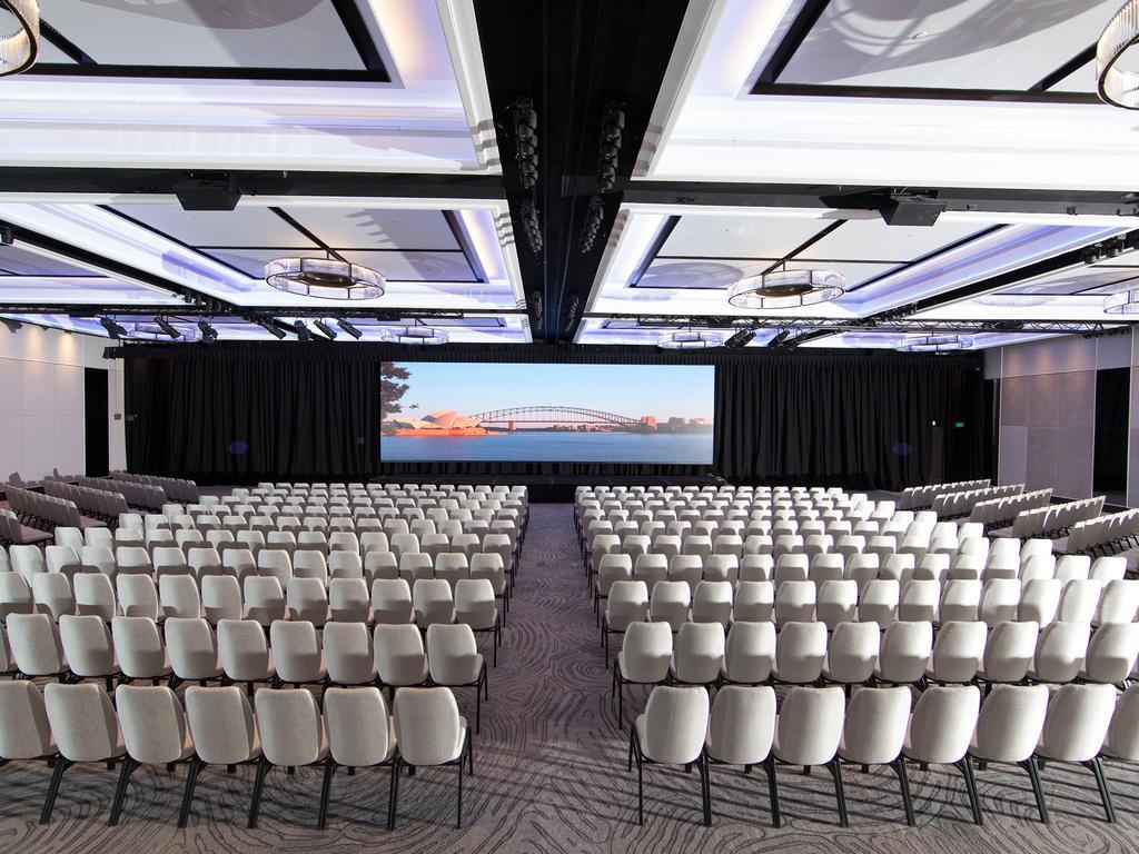 Four Seasons Hotel Sydney Esterno foto The photo depicts a large conference hall or auditorium filled with rows of white chairs arranged in a semi-circle facing a central stage or screen. The room has a modern design, featuring stylish ceiling lights and a sleek, contemporary aesthetic. A