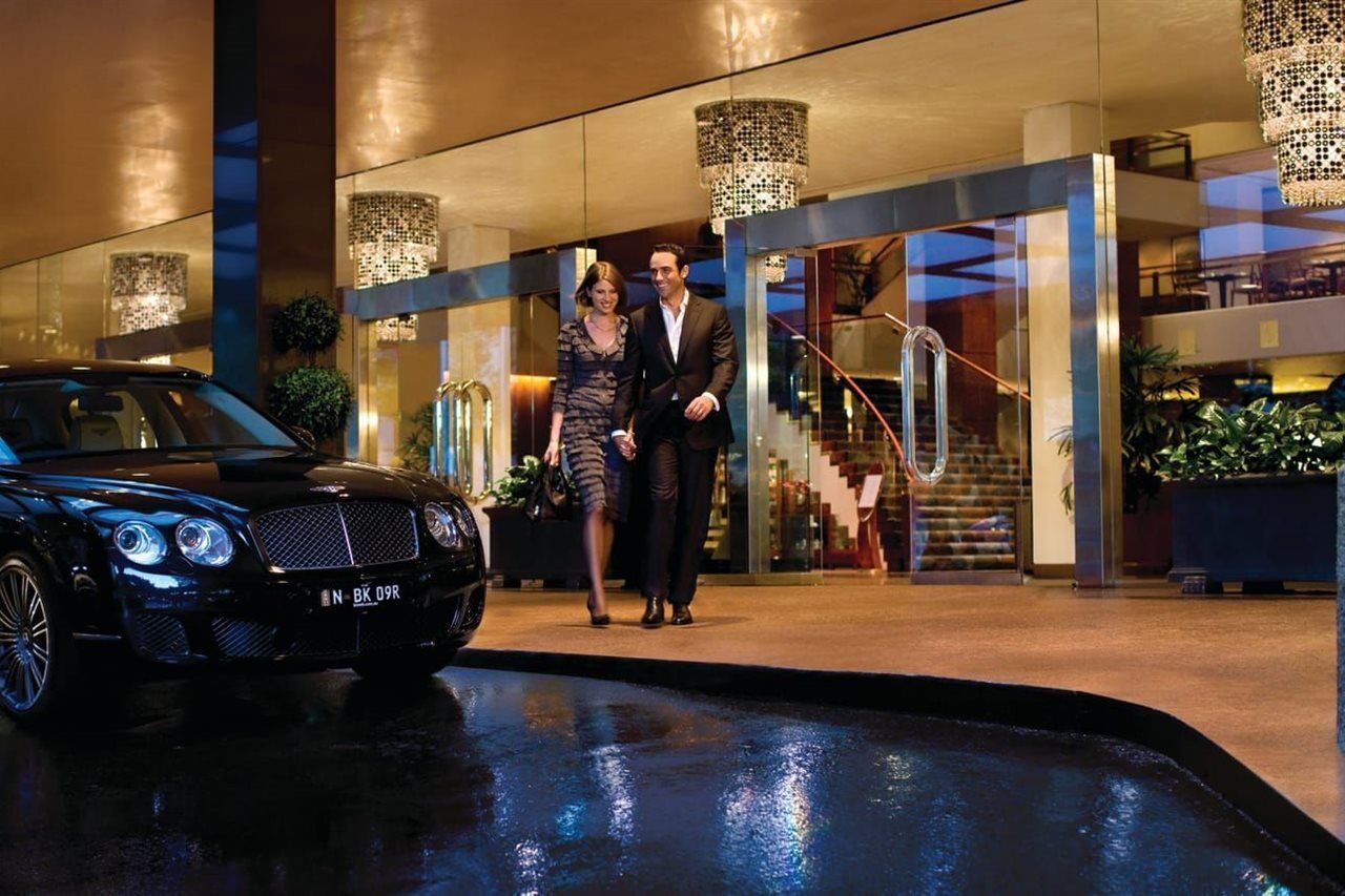 Four Seasons Hotel Sydney Esterno foto The photo shows a stylish couple walking together outside a luxurious hotel. The man is dressed in a formal suit, while the woman is in an elegant, patterned dress. They are holding hands and appear to be enjoying each other's company. In the foregro