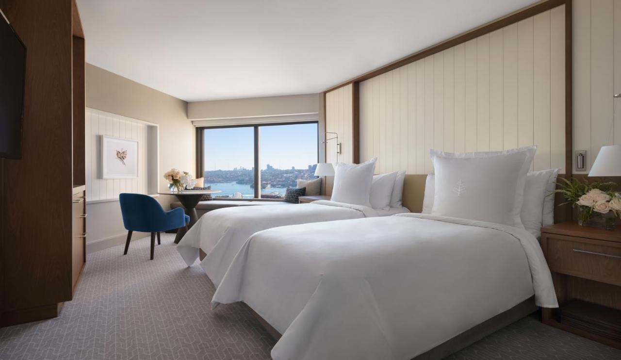 Four Seasons Hotel Sydney Esterno foto The photo shows a contemporary hotel room featuring two neatly made beds with white linens. There is a cozy blue chair positioned near a table, and a vase with flowers on the table adds a touch of decor. The large window offers a view of the outside,