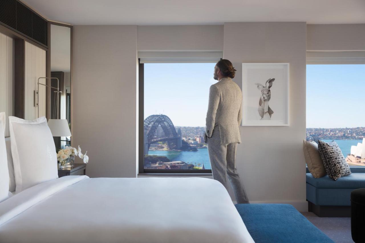 Four Seasons Hotel Sydney Esterno foto The photo features a modern hotel room with a large bed and a stylish decor. A person dressed in a formal outfit stands near a window, looking out. The view includes a scenic landscape, possibly of Sydney, showcasing the harbor and a landmark structu