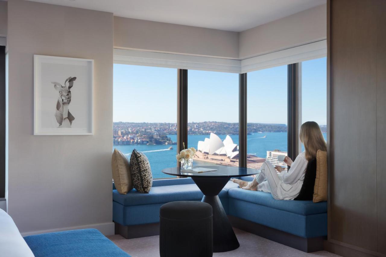 Four Seasons Hotel Sydney Esterno foto The photo shows a modern hotel room with a stunning view. A person is seated on a blue bench in front of large windows, gazing out at the scenery. There’s a round black table in the center, and the background features water and architecture, possibly